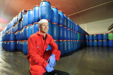 Mann med hårnett, hansker og rød kjeldress sitter på kne foran store stabler med marinert sild på tønner